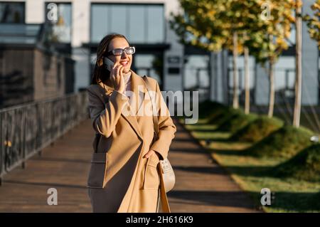 Stylish young woman in camel color autumn coat and sunglass walking on the street and talking on the smartphone, smiling lady enjoying phone conversation outdoors Stock Photo