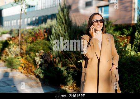 Cheerful young woman walking on the street chatting on the smartphone, female in elegant autumn coat enjoying phone conversation outdoor Stock Photo
