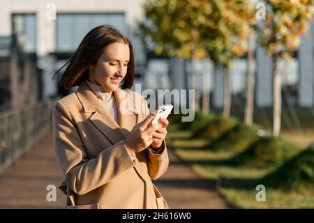 Young woman in camel color autumn coat walking on the street and using smartphone, smiling lady enjoying phone conversation online standing outdoors, texting Stock Photo