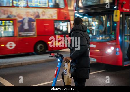 LONDON - NOVEMBER 1, 2021: A man holding a smartphone and riding an electric scooter with red double decker buses in the background at night Stock Photo