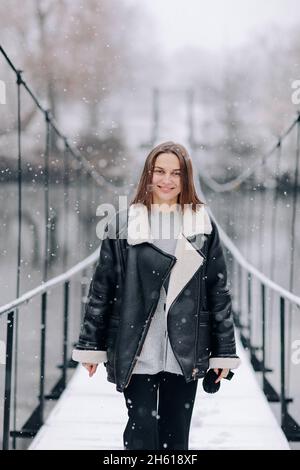 A woman walks over the river on a suspension bridge in winter day. Young girl in warm clothes stands on a wooden footbridge in cold snowy day. Stock Photo