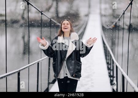 A woman walks and catches snowflakes over the river on a suspension bridge in winter day. Young girl in warm clothes stands on a wooden footbridge in Stock Photo