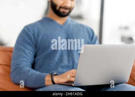 Cropped picture of Indian bearded man using laptop, Hispanic guy is typing on the keyboard, answering emails, multiracial male freelancer in casual wear working remotely from home Stock Photo
