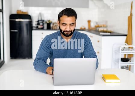 Focused handsome young Indian man is studying online, watching online webinar, video classes on the laptop. A clever multiracial man e-studying, doing researching sitting at home Stock Photo
