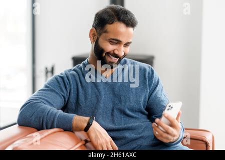 Handsome Indian guy using a smartphone for video call, smiling man in casual wear talking into webcam, enjoying virtual meeting with friend or girlfriend sitting on the couch at home Stock Photo