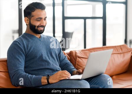 Cheerful Indian man wearing casual wear sitting on the sofa and using laptop at modern home office, glad ethnic latin male entrepreneur typing on keyboard Stock Photo