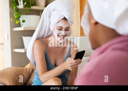 Happy woman with facial mask using smartphone while sitting with female friend at home Stock Photo