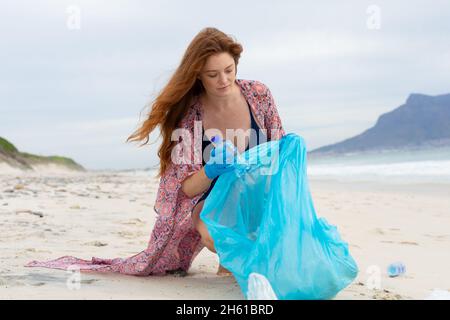 Caucasian young woman in casuals collecting plastic waste in bag at beach Stock Photo