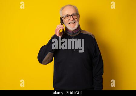 Lifestyle, technology and old people concept: Handsome old gray-haired man dressed in smart casual style using a smartphone and smiling over yellow ba Stock Photo