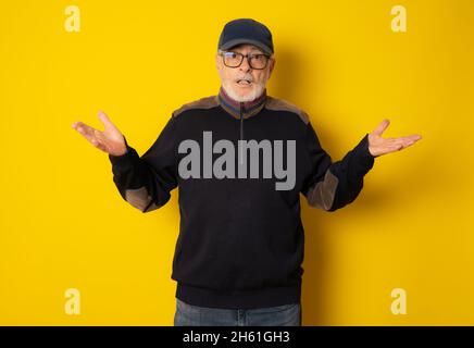 Worried mature man in winter clothing and cap with open arms gesture isolated over yellow background. Stock Photo