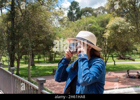 Beautiful Hispanic woman with a hat taking a picture with an old camera in the middle of a park on a sunny morning Stock Photo