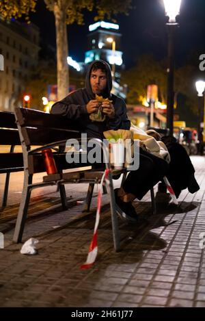 Homeless guy in hoodie chewing burger and looking away while sitting on bench on illuminated street at night Stock Photo