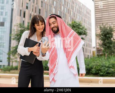 Two smiling diverse business people arabian man and caucasian woman standing outdoors on city street and using smartphone, arab man showing something on mobile phone to his female assistant Stock Photo