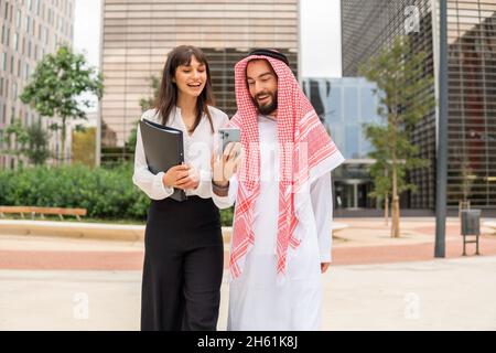 Smiling Arabian businessman in traditional wear and his female assistant secretary watching funny videos on smartphone, standing on city street with skyscrapers on background Stock Photo