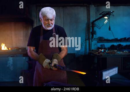 Caucasian aged man in safety apron and gloves working with molten metal at forge. Industrial worker with grey beard making some metal product at work.  Stock Photo