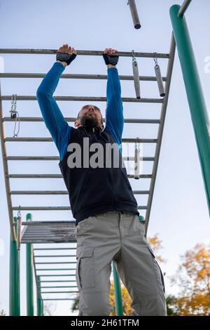 Strong Grey-Haired Man With Dreadlocks rearranges his hands on horizontal monkey bar outdoor. Climbing frame for sport activity in open air. Adult Stock Photo