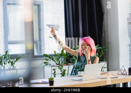 happy businesswoman with pink hair talking on smartphone while throwing paper plane Stock Photo