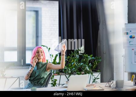 positive businesswoman with pink hair talking on smartphone while throwing paper plane Stock Photo