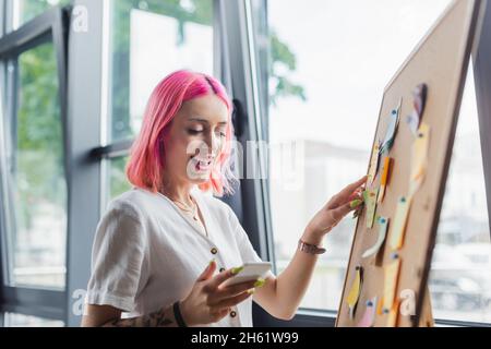 happy businesswoman with pink hair holding smartphone and pointing at board with sticky notes Stock Photo