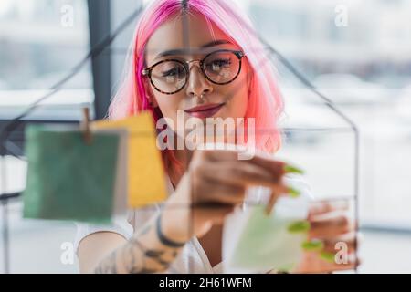 smiling businesswoman with pink hair holding wooden pin near blurred sticking note on metallic board Stock Photo