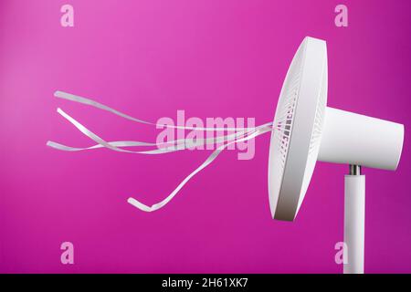 The electric fan is white with pink ribbons fluttering in the wind on a pink background. Free space, minimalistic style. Stock Photo