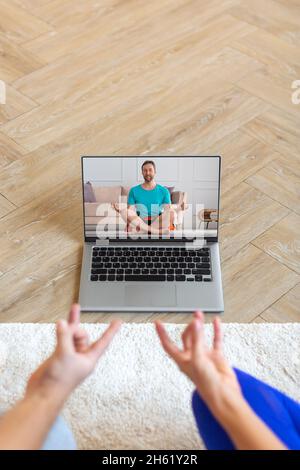 A couple meditates under the guidance of an online trainer in front of a laptop monitor. The concept of mental health and yoga practices using modern Stock Photo