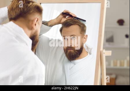 Confident handsome bearded young man looking in bathroom mirror and brushing his hair Stock Photo