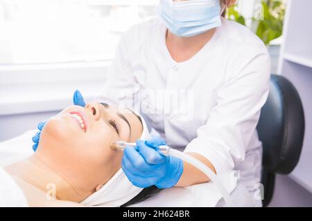 Cosmetologist in a protective medical mask and gloves making the facial microdermabrasion rejuvenation procedures to her client in a beauty salon. Stock Photo