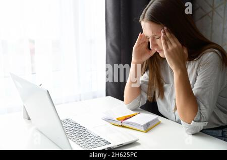 Tired and worn out from online work young woman sitting in front laptop holding head, feels strong headache, female freelance with eyes closed is massaging head temples with suffering face expression Stock Photo