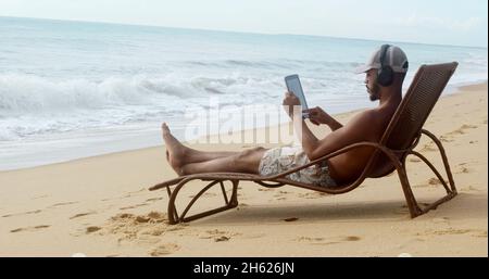 Man making a video call on summer vacation. latin american man sitting on the beach chair with headphones and a tablet Stock Photo