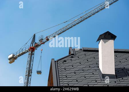 Construction site with crane and a closeup of a roof of a residential building with a chimney on a clear blue sky. Construction industry concept. Stock Photo