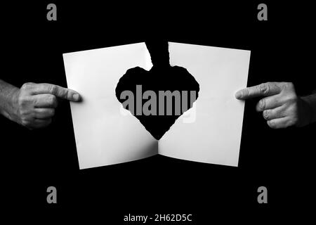 B+W image of male hands holding and ripping sheet of paper with torn heart shape against black background. Stock Photo