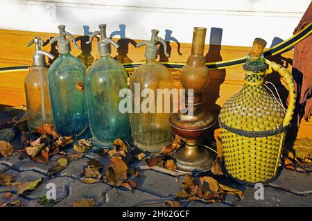 Four old glass siphons, a kerosene lamp and a glass bottle for wine in a plastic braid among the dry fallen leaves on the pavement Stock Photo