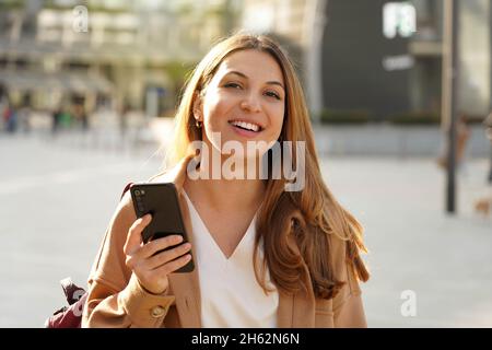 Portrait of cheerful multi ethnic female in trendy wear spending time on street using smartphone, beautiful millennial girl with coat looking at camer Stock Photo