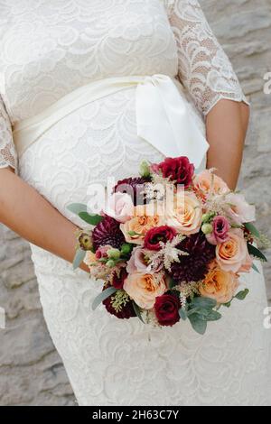 pregnant bride in a white lace dress with a colorful bridal bouquet in front of her belly Stock Photo