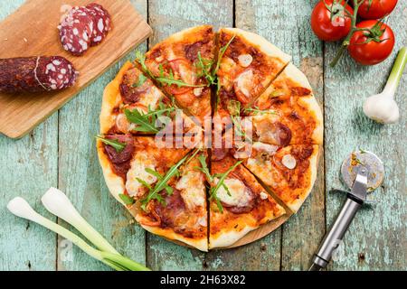 Pizza with fresh rocket salad cut in segments. Cherry tomatoes, salami, garlic, leek and pizza knife on background overhead view Stock Photo