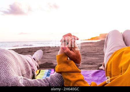close up of two hands holding together at the beach with the sunset at the background enjoying summer and having fun together. couple of people on the sand Stock Photo