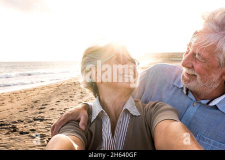 portrait of couple of mature and old people enjoying summer at the beach looking to the camera taking a selfie together with the sunset at the background. two active seniors traveling outdoors. Stock Photo