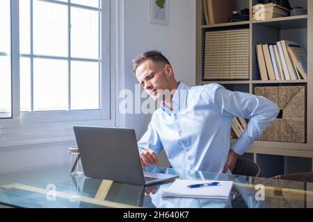 close up rear view stressed young man touching lower back feeling discomfort,suffering from sudden pain due to sedentary lifestyle or long computer overwork in incorrect posture at home office. Stock Photo