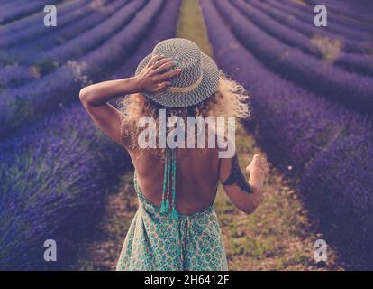back view of pretty babe in boho elegant blue dress walk in lavender fields wearing travel style hat and blonde curly nice long hair - concept or free woman in the outdoor nature adventure lifestyle