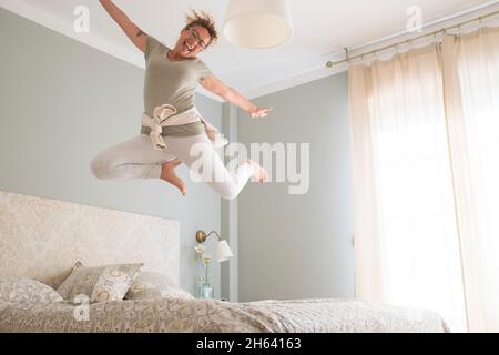 young adult pretty woman go crazy for happiness and jump on the bed with coffee cup - overjoyed female caucasian people alone at home have fun jumping in bedroom with craziness Stock Photo