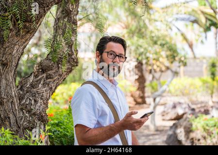 portrait of businessman using cellphone outdoor at the park with roaming internet connection - adult bearded man with glasses looking on camera Stock Photo