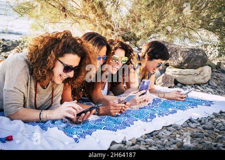 group of young adult women use modern phones together having fun and laughing a lot - happy females people enjoying technology and internet connection outdoor laydown Stock Photo