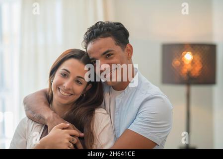 young millennial interracial couple hug with love at home in living room - relationship with black boy and caucasian girl together standing and embracing looking each other - concept of life and house Stock Photo