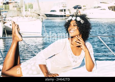 happy african american woman with curly hair lying down barefoot on private yacht. joyful black woman in white dress having fun on boat. female tourist posing on nautical vessel Stock Photo