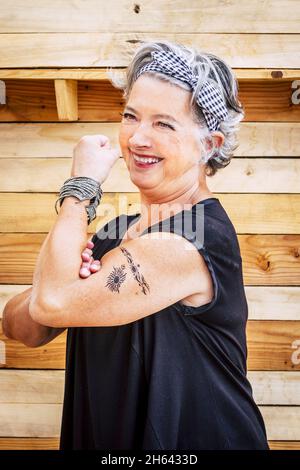 senior man in t shirt and bandanna showing biceps. cheerful elderly woman posing in front of wooden wall. tattooed old woman having fun flexing her biceps Stock Photo