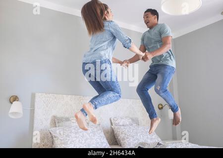 overjoyed young couple holding hands and jumping on bed. beautiful couple jumping midair on bed. carefree couple holding hands while dancing and jumping on bed in modern apartment Stock Photo