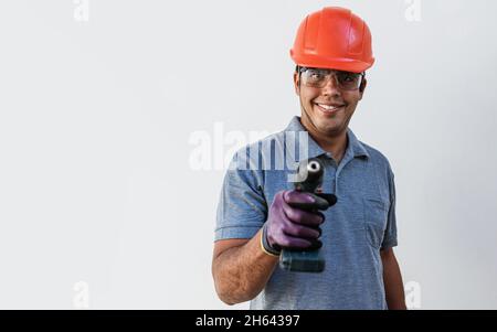Confident young Latin male carpenter in hardhat holding drill and looking at camera with smile while standing against white background. Always ready t Stock Photo