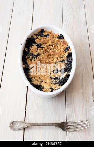Blueberry crumble pie in oval white cooking pan with blueberries and old fork on bright wooden background high angle view Stock Photo