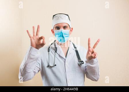 Present doctor in white suit and protective breathing mask glasses and stethoscope showing peace signs V and OK by his hands Stock Photo
